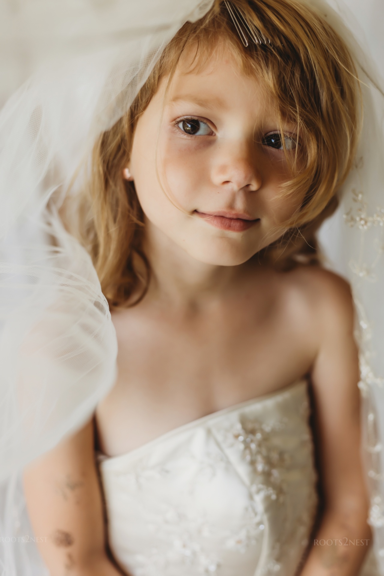 Young girl with light brown hair and big eyes, looking at the camera, wearing a white dress, surrounded by soft fabric.