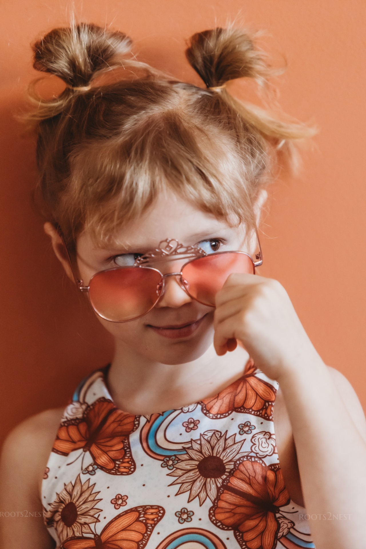 Young girl with two buns, wearing sunglasses and a floral-patterned shirt, smiling and touching her nose, against a peach background.