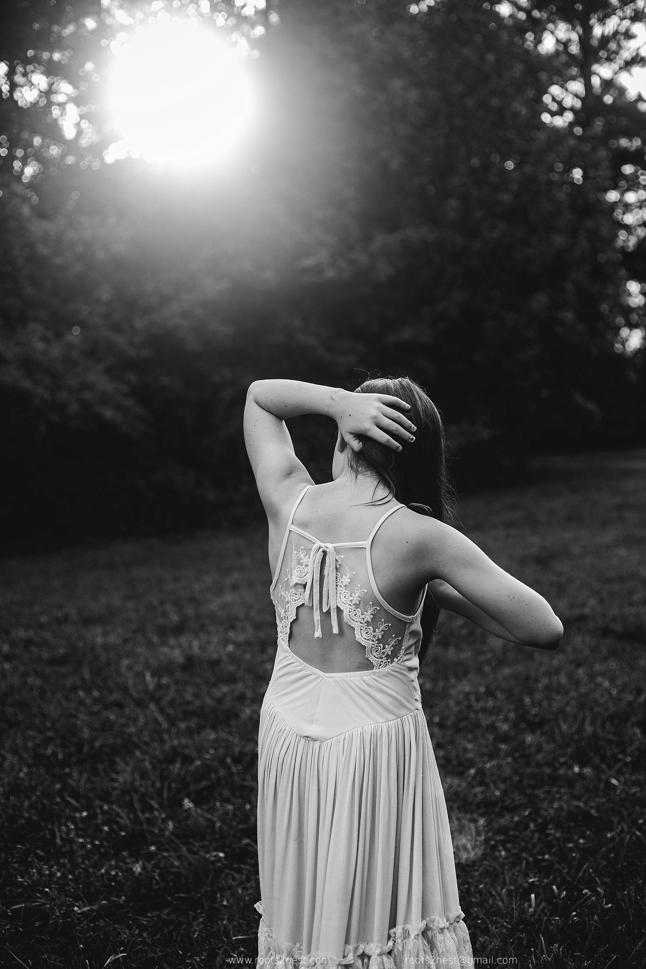 Woman in a flowing dress stands outdoors with her back to the camera, hand on her head, sunlight through trees behind her.