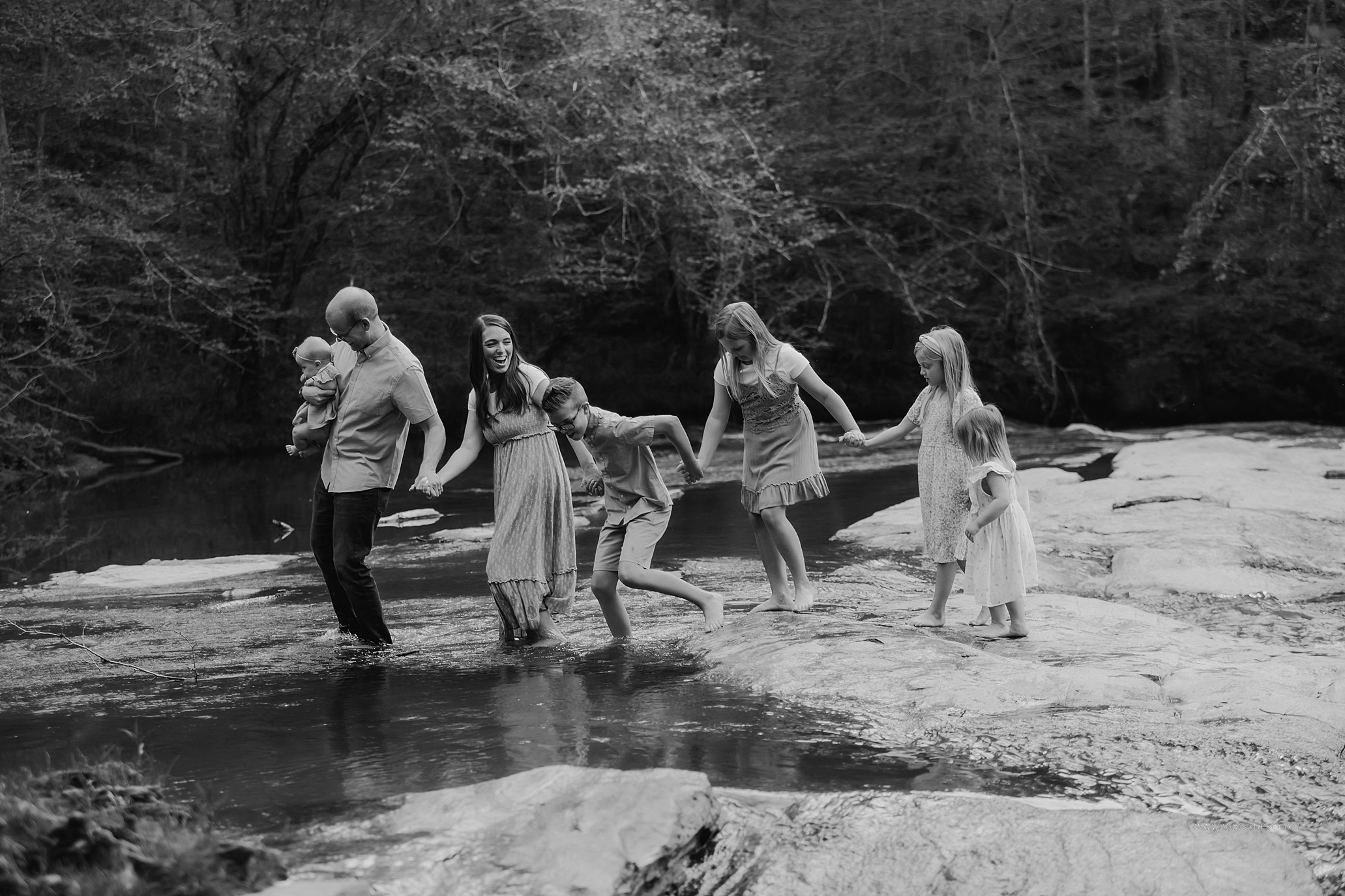 Group of six people holding hands and walking across a shallow river in a natural setting.