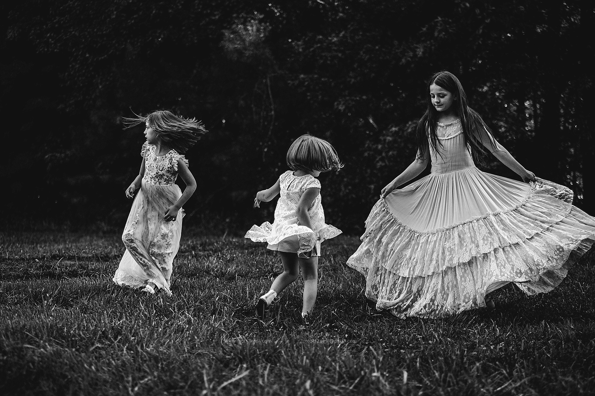 Three girls playing outdoors on grass, one in a long dress, two in shorter dresses, trees in background.