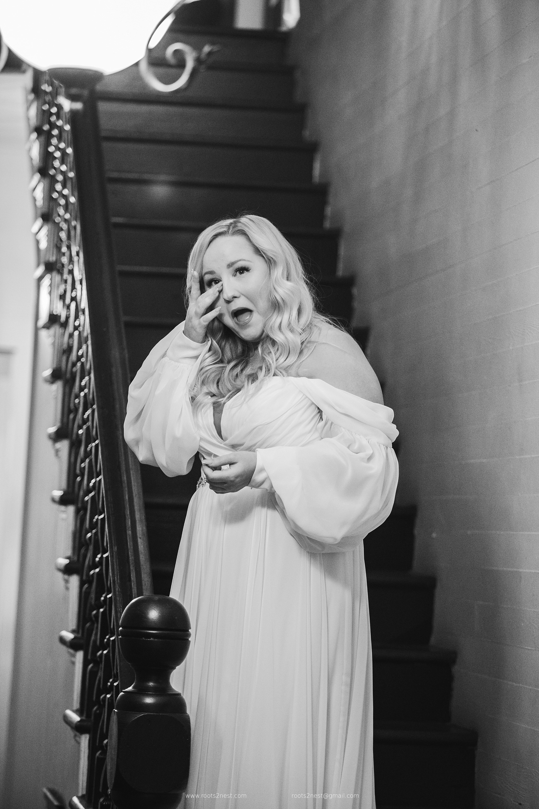 Woman with wavy hair in a long dress standing on a staircase, holding her hand to her face, smiling.