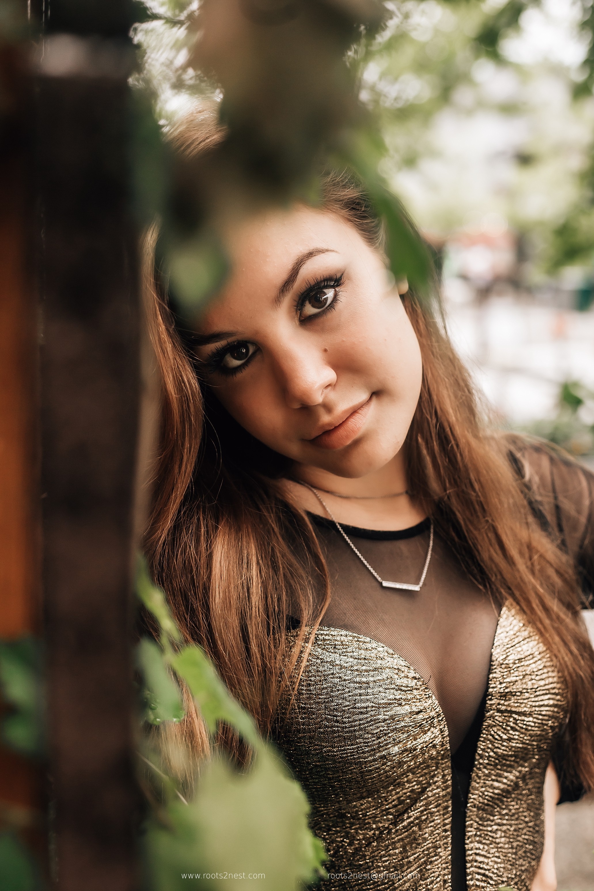 Young woman with long brown hair and makeup, looking at camera outdoors, wearing a black top with a gold-patterned bust, surrounded by greenery.