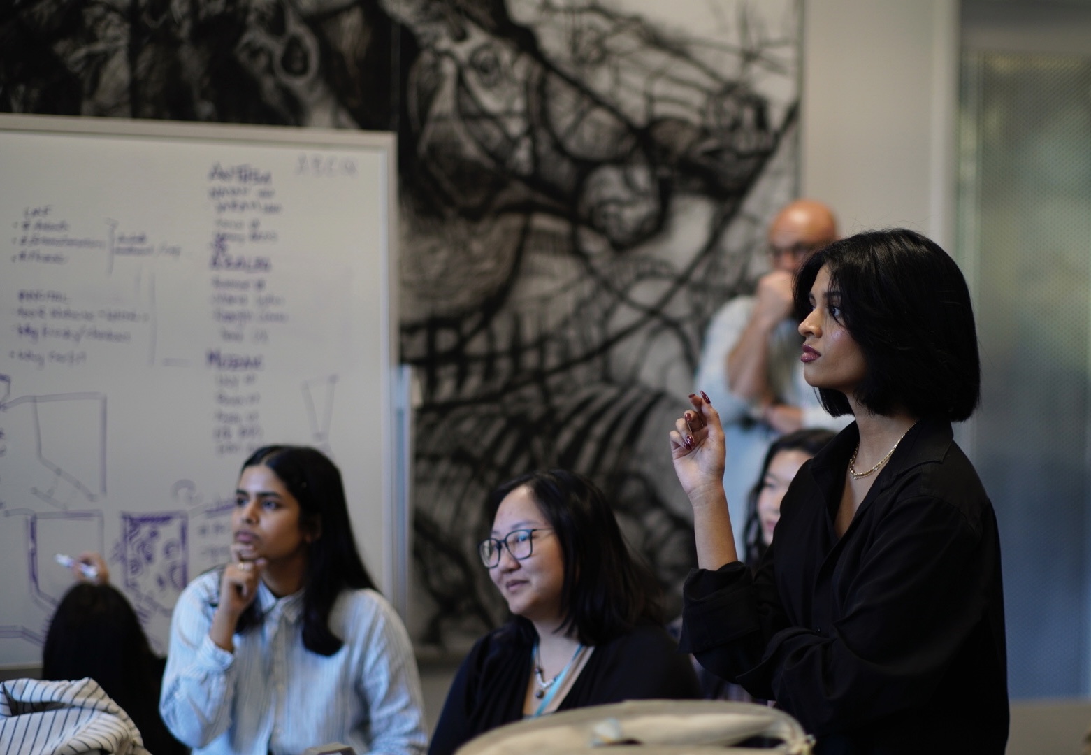 Three women seated and one woman standing in a room with a whiteboard and abstract wall art, engaging in discussion.