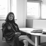 Woman sitting at a desk near windows, smiling, with a laptop and papers, in a bright room.