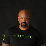 Man with a shaved head and goatee wearing a black T-shirt with yellow text, against a dark background.