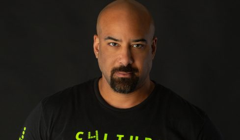 Man with a shaved head and goatee wearing a black T-shirt with yellow text, against a dark background.