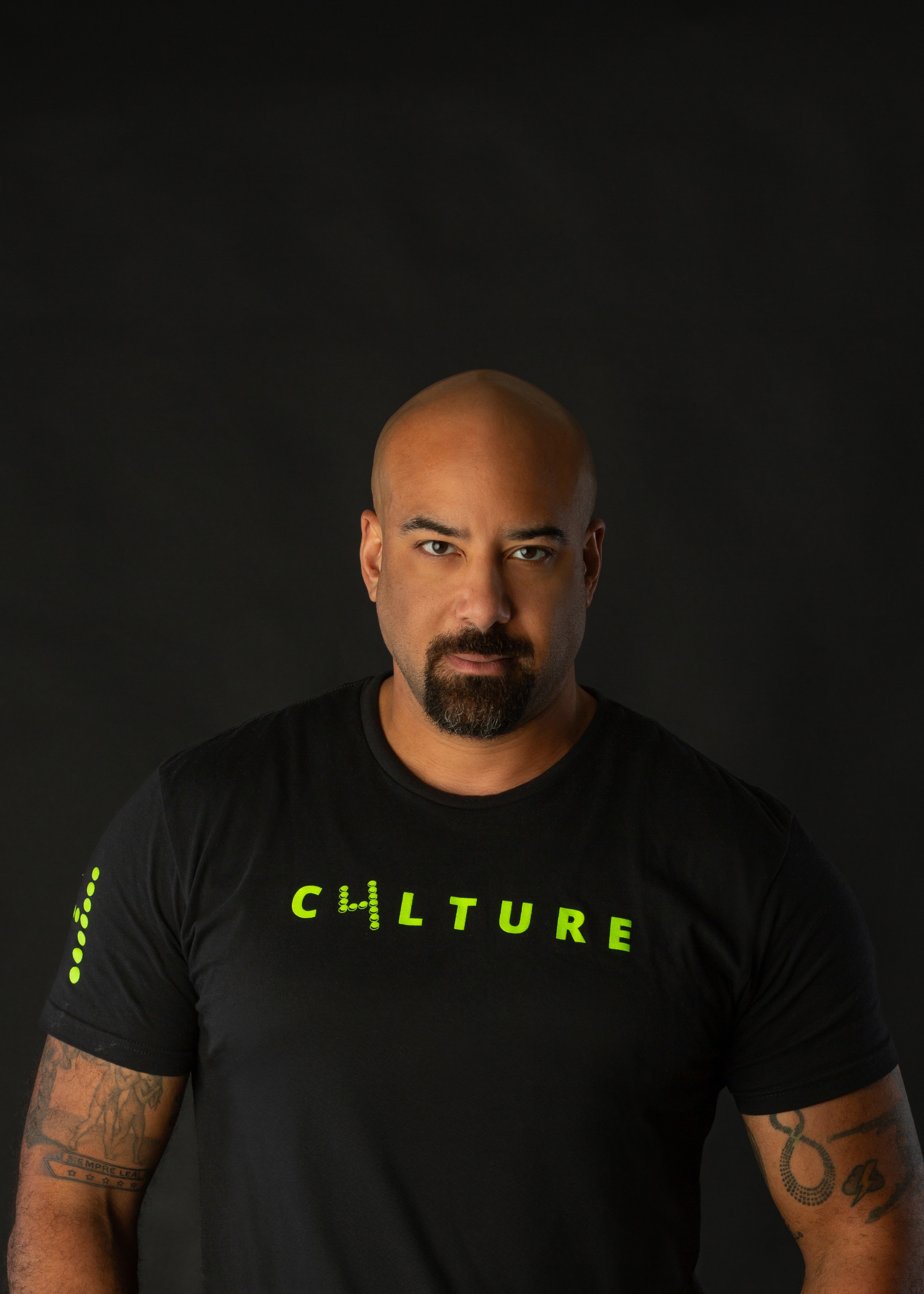 Man with a shaved head and goatee wearing a black T-shirt with yellow text, against a dark background.