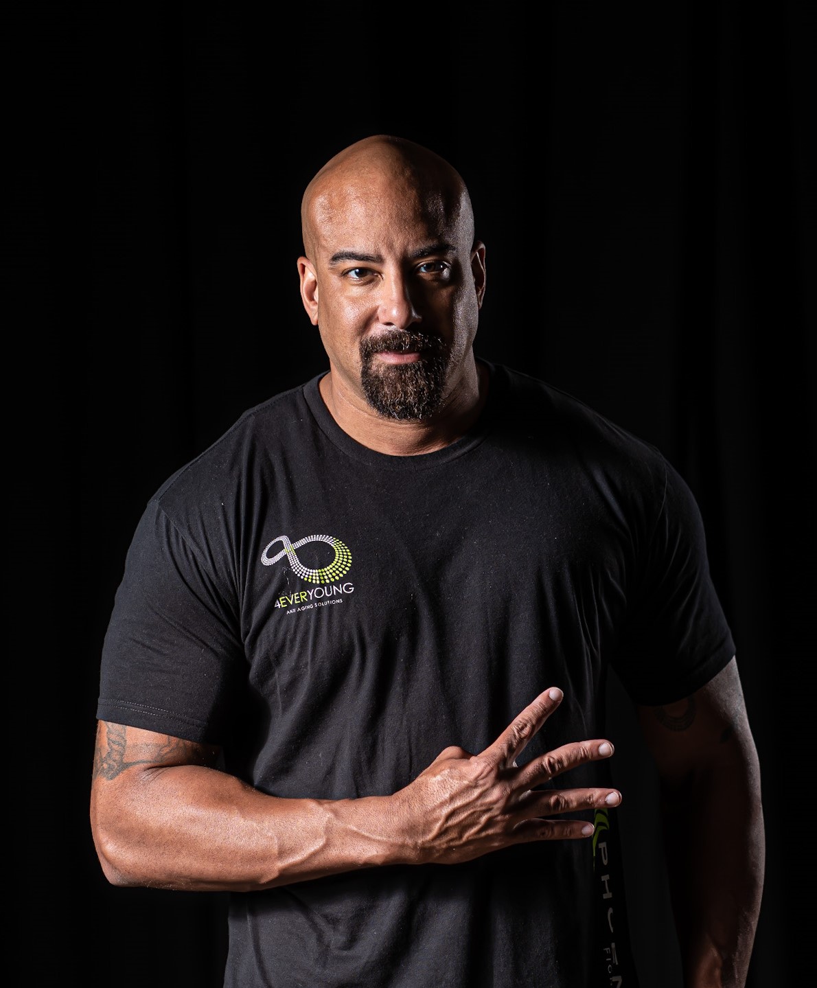 Man with a beard and shaved head wearing a black T-shirt making a peace sign gesture with his right hand, dark background.