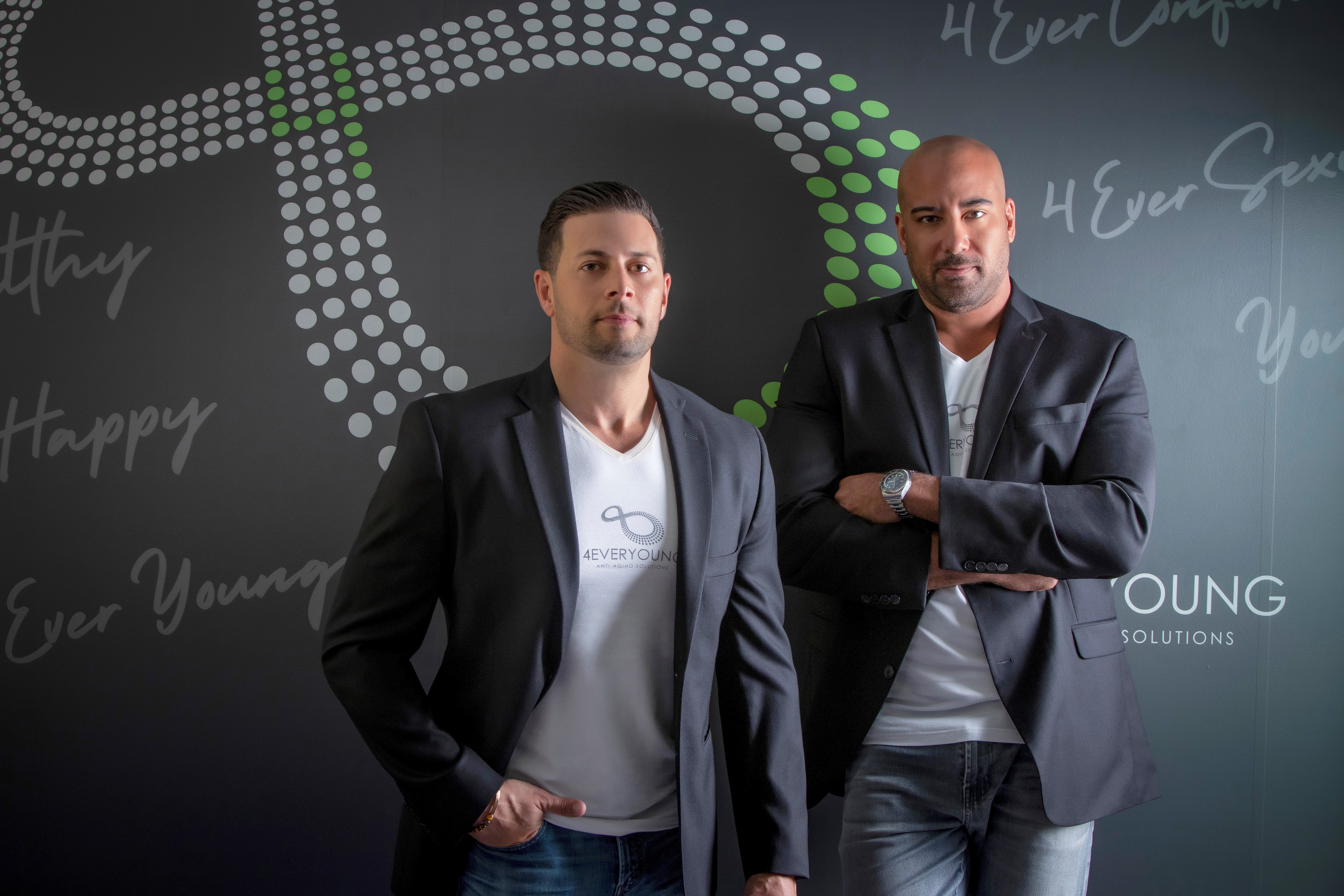 Two men in suits stand in front of a dark background with text and graphics, one with hands in pockets, the other with arms crossed.