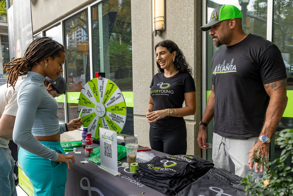 Three people at an outdoor promotional booth with a spinning wheel, drinks, and branded items, engaging in conversation.
