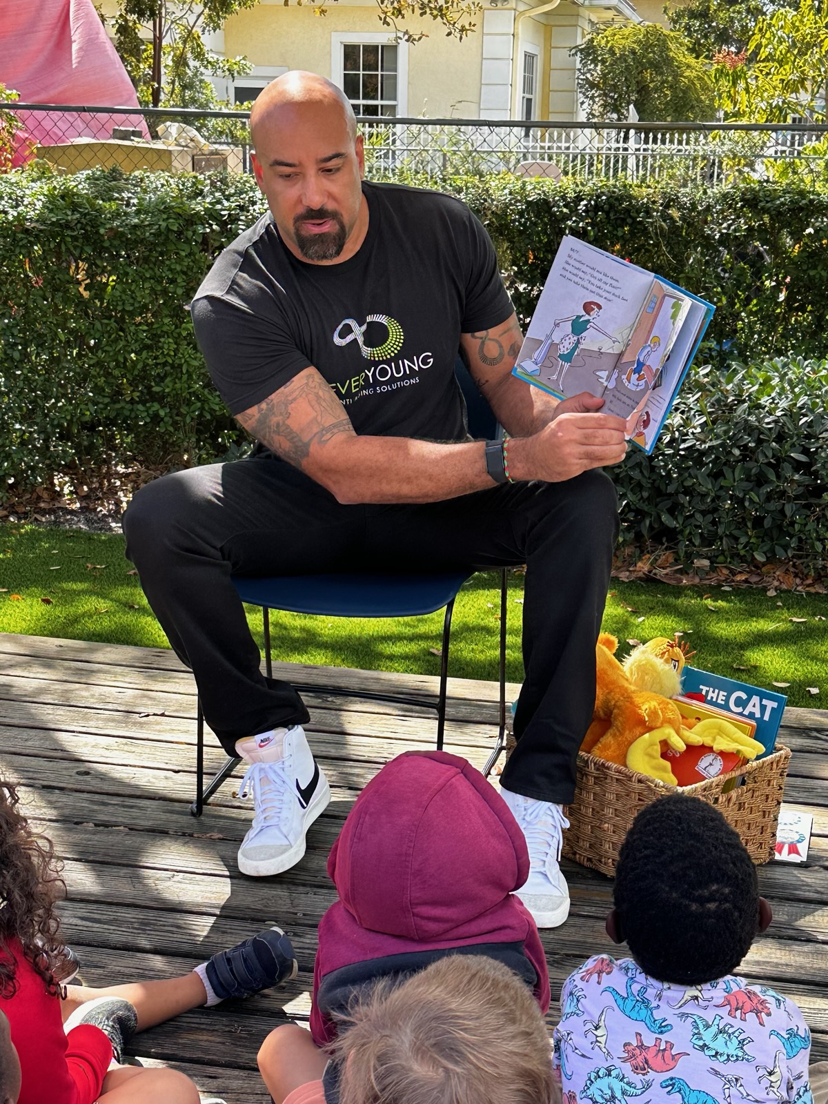Man sitting outdoors on a chair, reading a children's book to a group of children, with a basket of toys nearby.