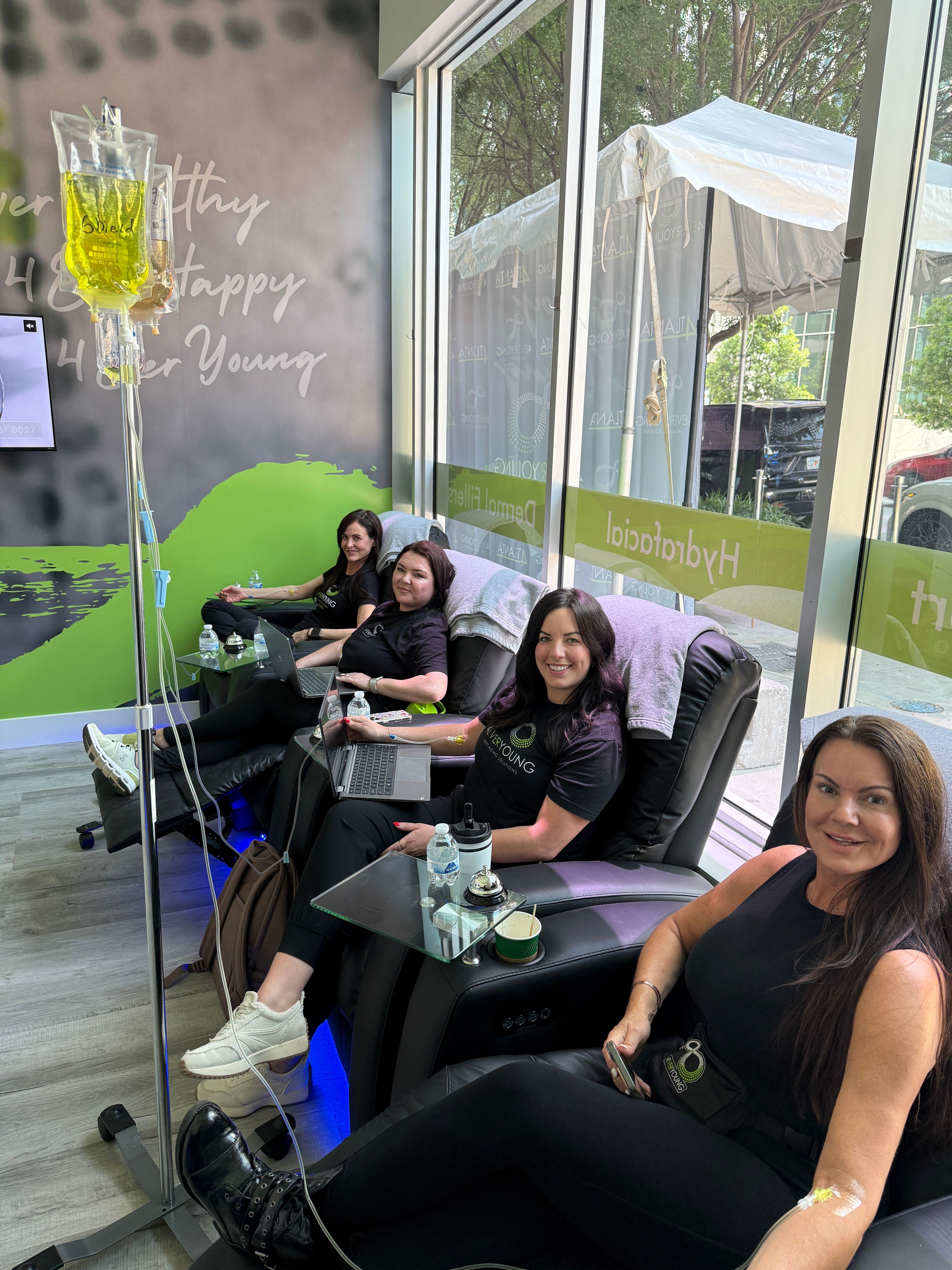 Four women sitting in a nail salon, smiling, with large windows and a green wall in the background.