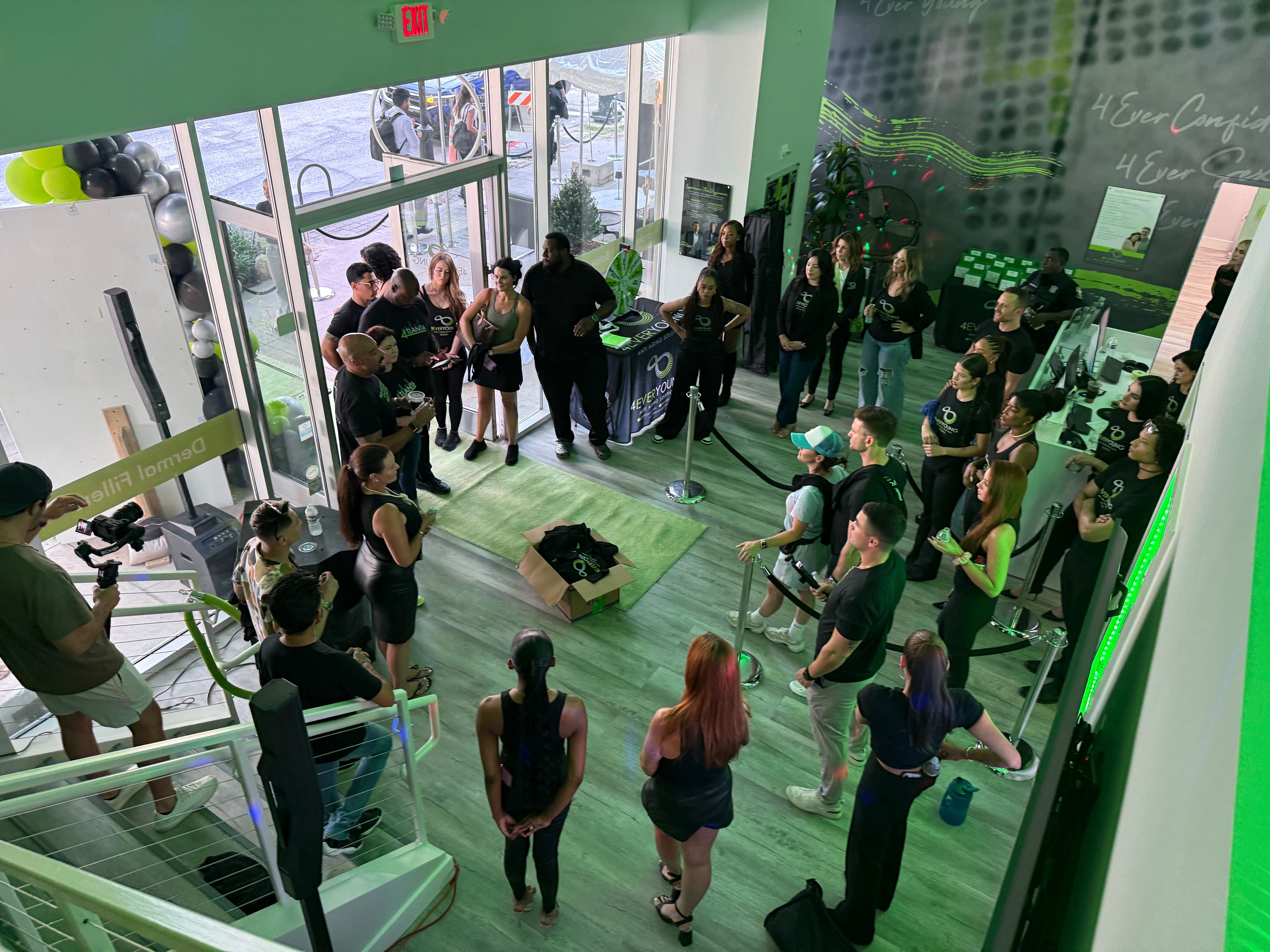 Group of people gathered around a green booth at an indoor event, some taking photos and listening to a speaker.